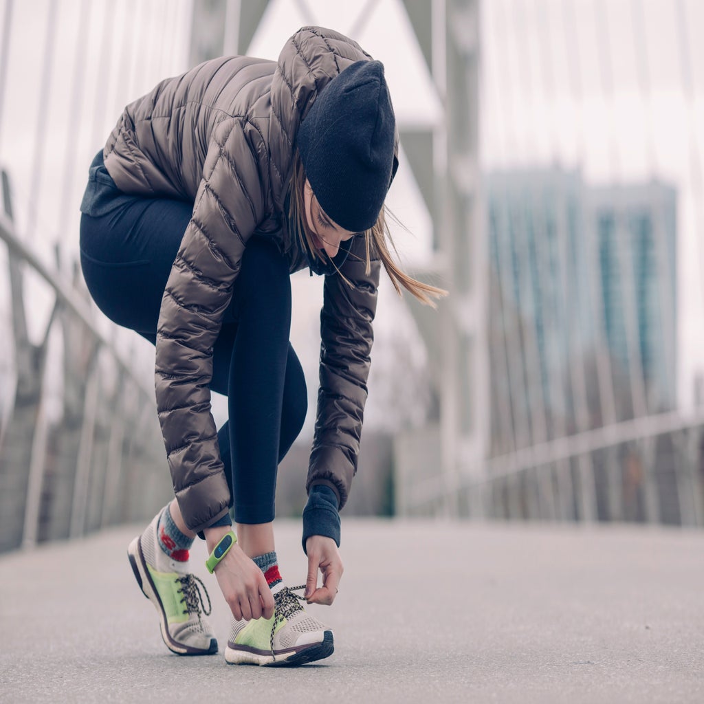 person kneels to tie their running shoes. they are wearing a coat and a hat and appear to be on a bridge