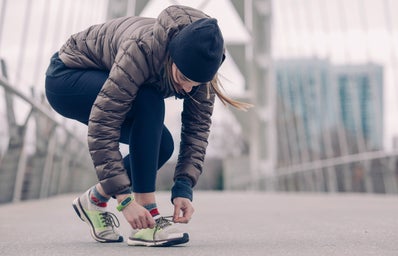 person kneels to tie their running shoes. they are wearing a coat and a hat and appear to be on a bridge