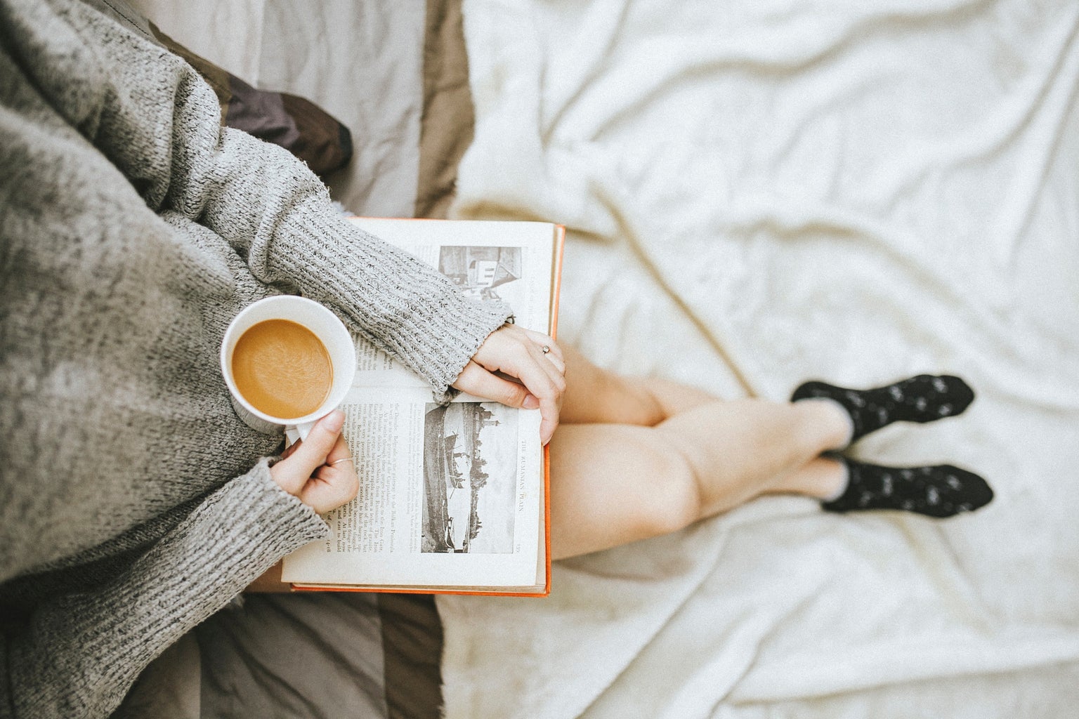person sitting at the edge of a bed with an open book in their lap and a cup of coffee in hand