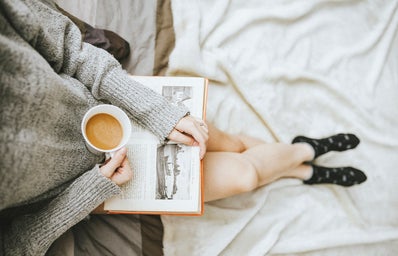 person sitting at the edge of a bed with an open book in their lap and a cup of coffee in hand