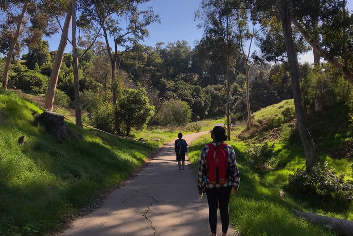 two people standing on a trail (spaced apart) with green trees all around them; the person closest to the camera has a red backpack