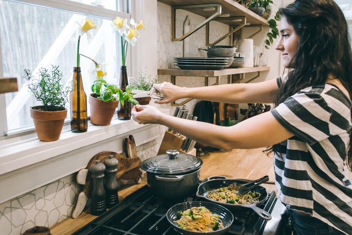a woman stands over the stove cutting an herb out of a pot on the window sill with a pair of scissors. there are pans of pasta stirfry on the burner.