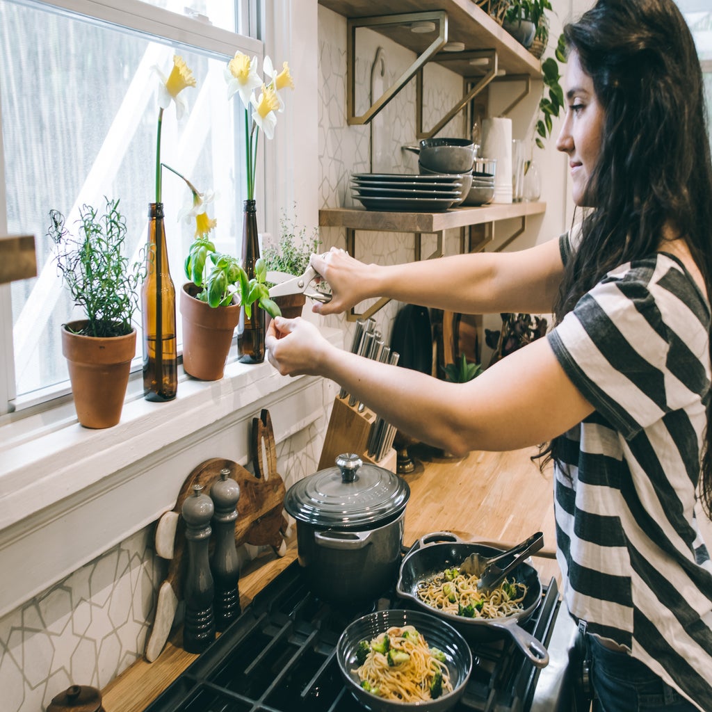 a woman stands over the stove cutting an herb out of a pot on the window sill with a pair of scissors. there are pans of pasta stirfry on the burner.