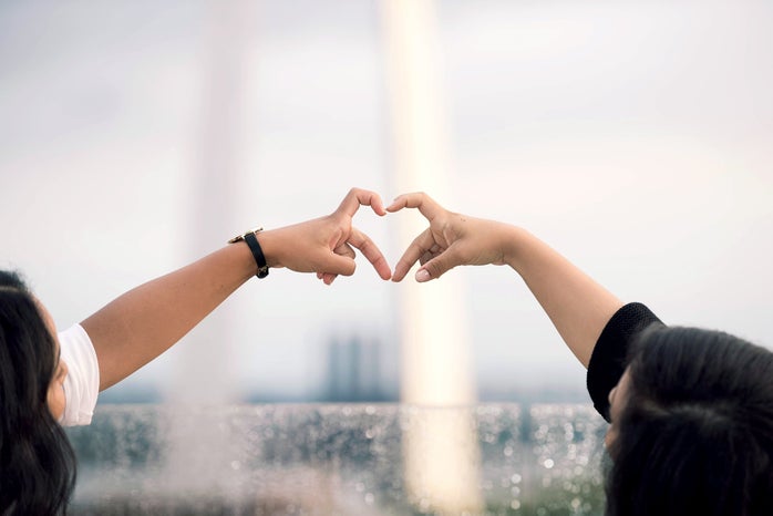 two different people's arms reach out in front of the St. Louis arch, their pointer finger and middle fingers coming together to make a heart