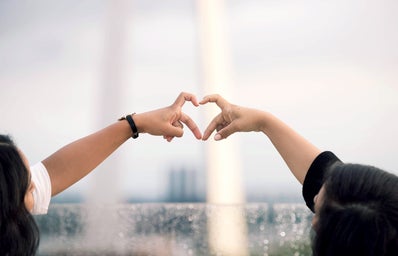 two different people\'s arms reach out in front of the St. Louis arch, their pointer finger and middle fingers coming together to make a heart