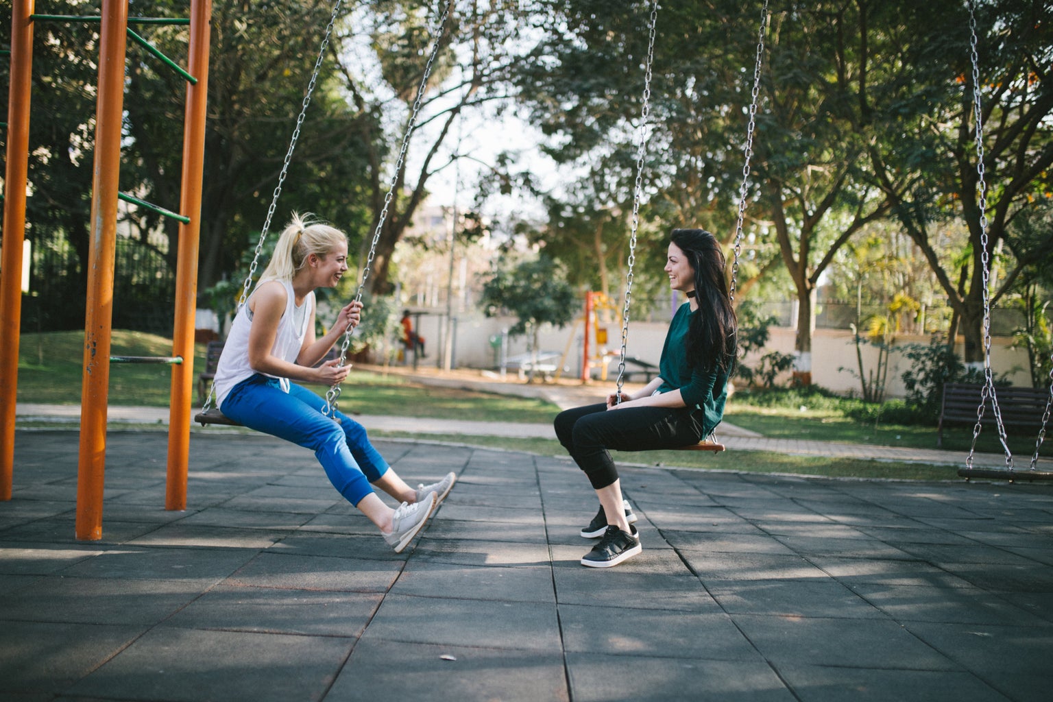 two women sit on a swing set. they are facing each other. 