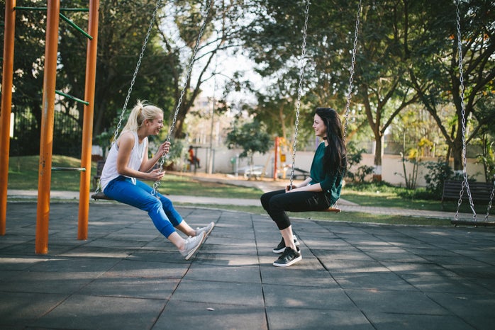 two women sit on a swing set. they are facing each other.