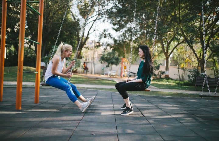 two women sit on a swing set. they are facing each other.