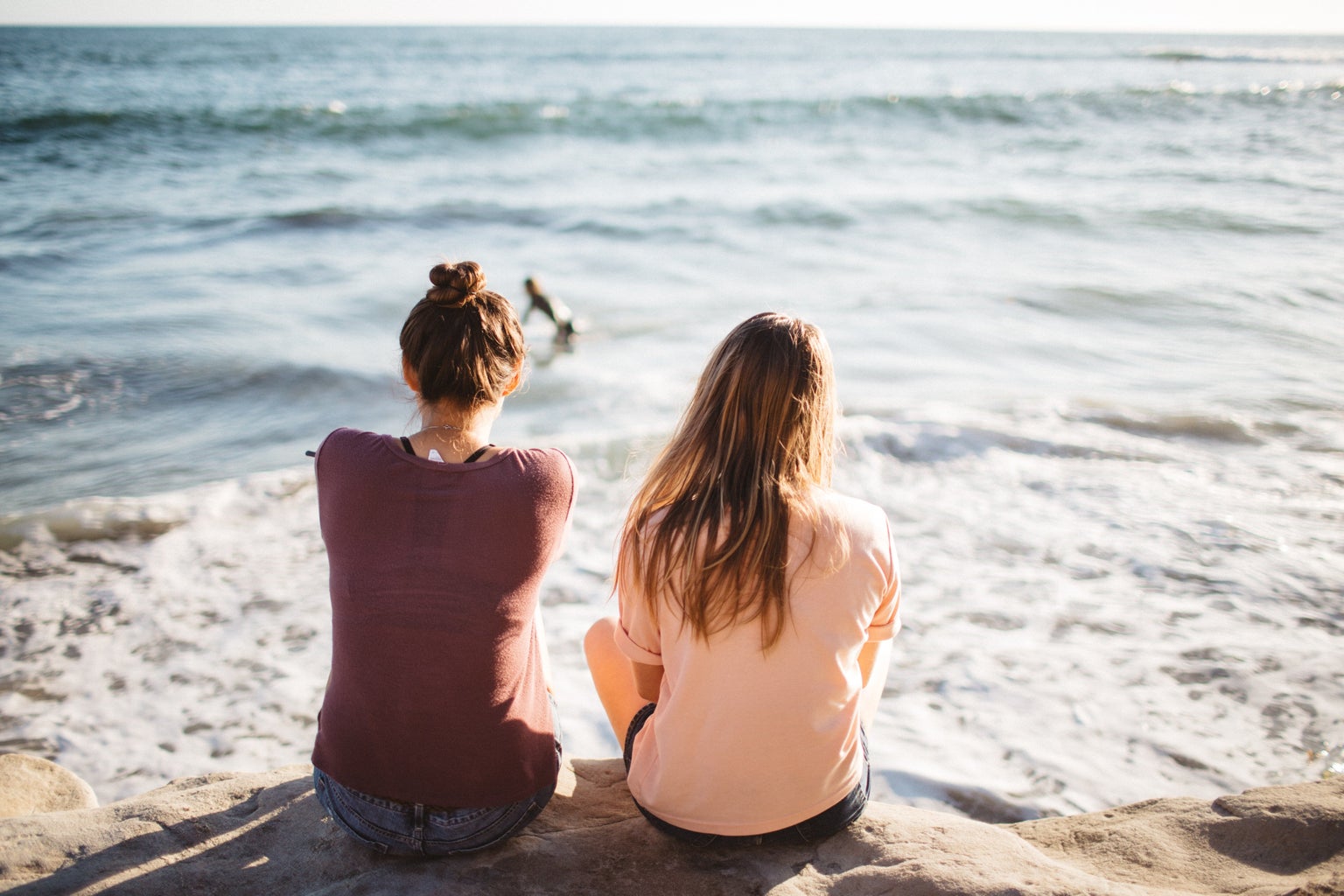 two women sit at the edge of the ocean facing the water