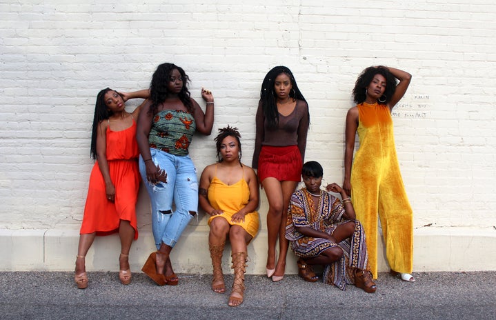 six black women standing and sitting agaisnt a white wall