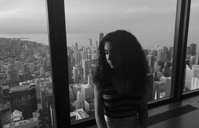 a B&W photo of a girl sitting on a window ledge with the skyline in the background