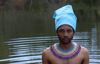 A Xhosa man, in traditional attire, stands infront of a body of water.