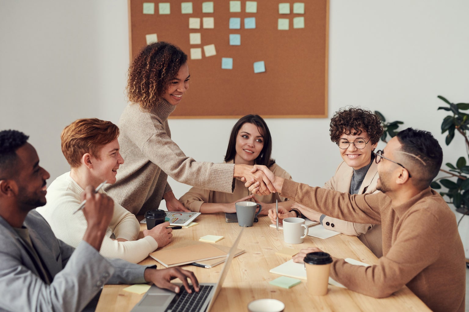 A group of people are in a meeting. They appear to be in a conference room at work. A woman is standing and shaking hands across the table with a man who is sitting down.