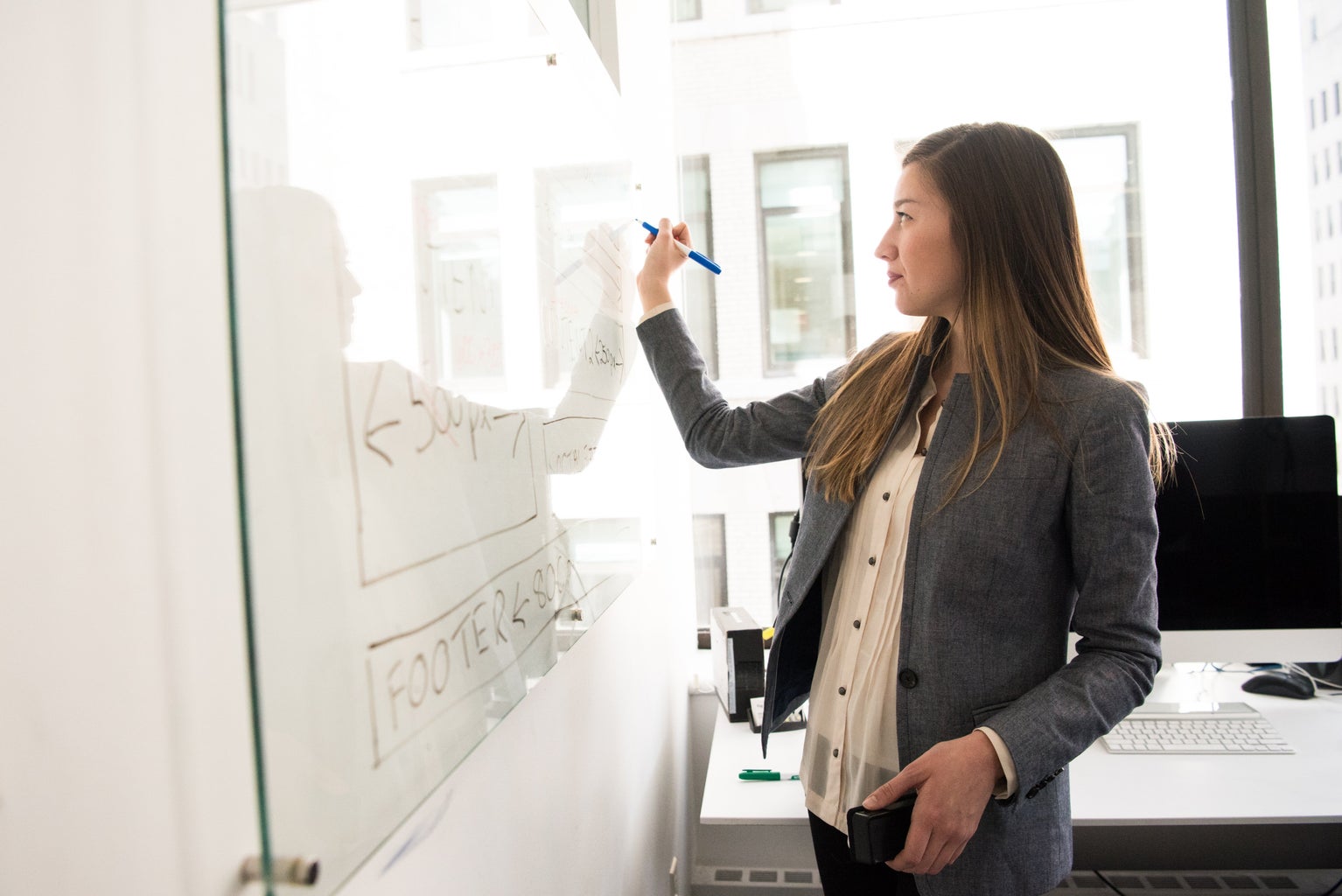 a woman in business casual stands in front of a white board, writing with a marker in an office space