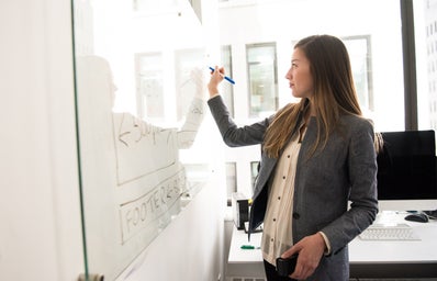 a woman in business casual stands in front of a white board, writing with a marker in an office space