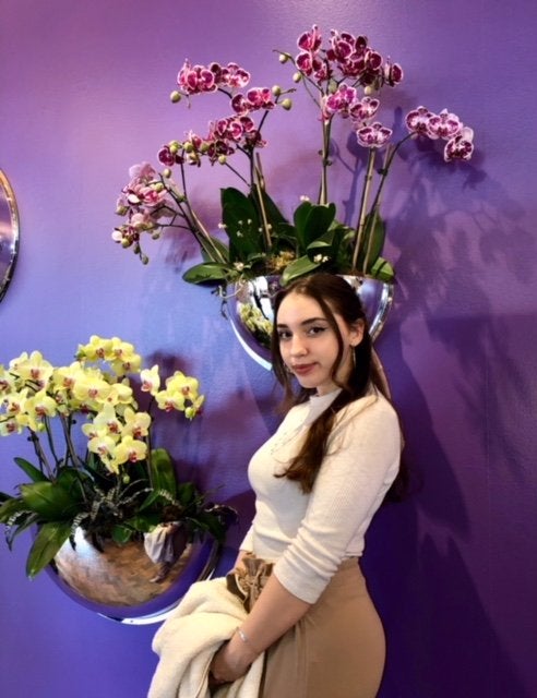 Girl smiling slightly, in front of purple and yellow orchids on purple wall