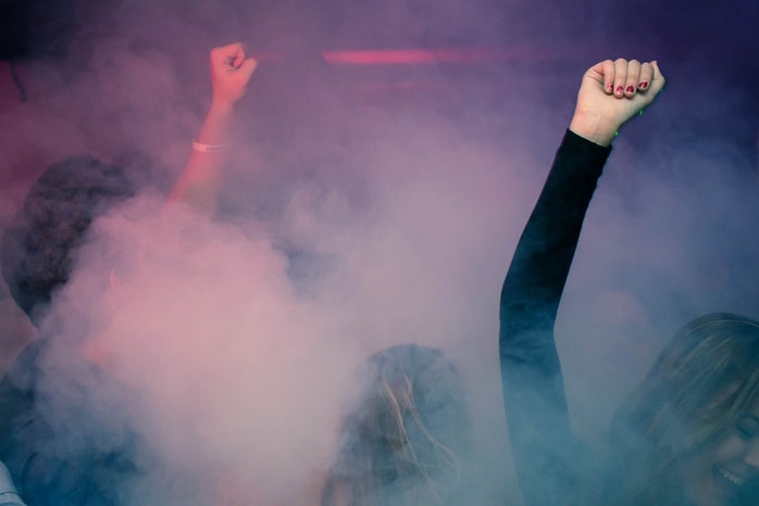 Women at a party with their arms raised. Purple and pink smoke in the background.