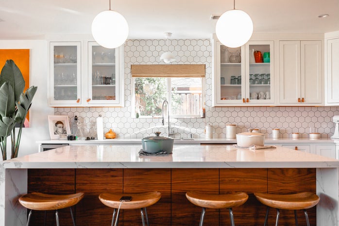 an island in a kitchen with a breakfast bar. there is a window over the sing and glass-fronted cabinets. the walls are white hexagonal tiles and there is an orange accent