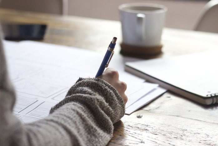 a hand holds a pen writing on sheets of paper on a wooden desk. there's a coffee cup and a notebook in front of it.