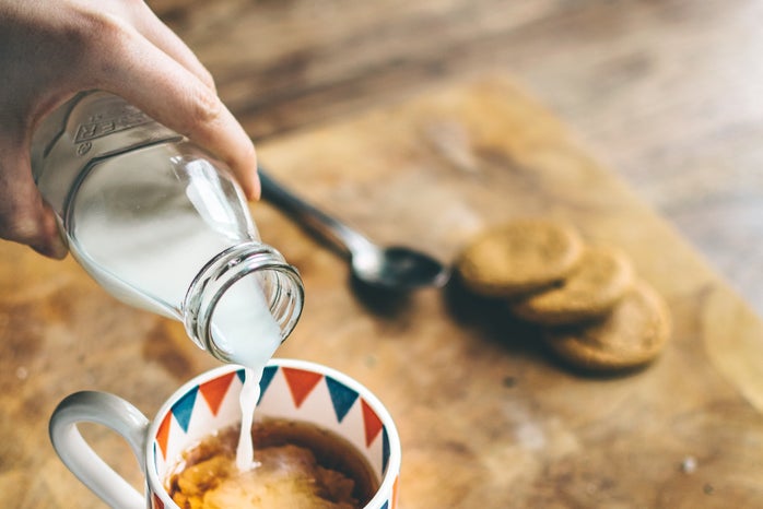a hand is pouring milk out of a glass bottle into a cup of coffee, which is sitting on a wooden cutting board on a wooden table.