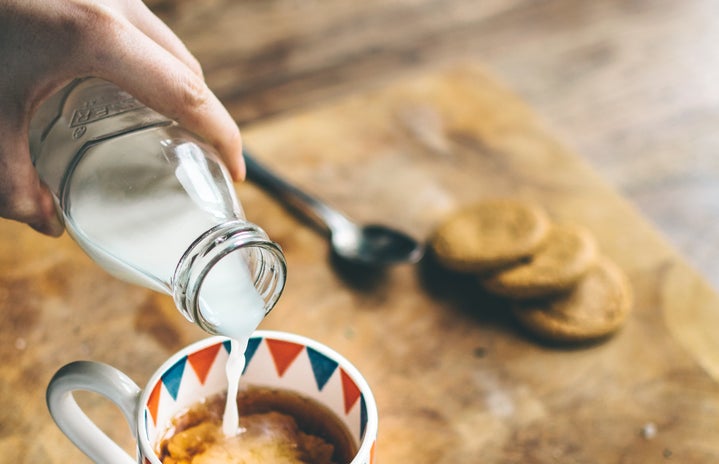 a hand is pouring milk out of a glass bottle into a cup of coffee, which is sitting on a wooden cutting board on a wooden table.