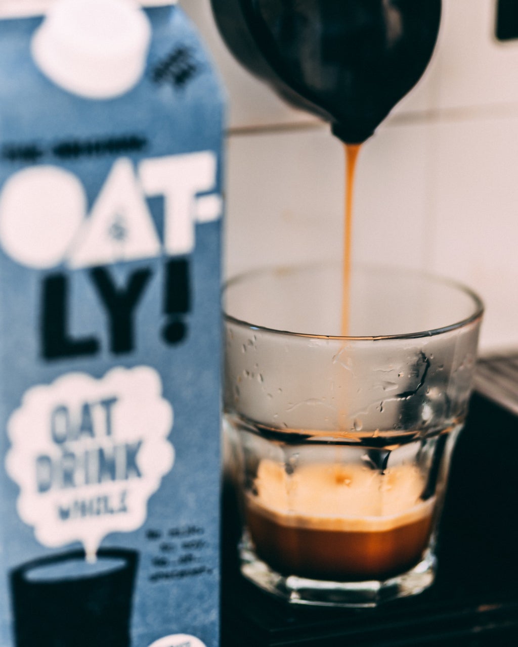 coffee is poured into a glass cup on a counter. there is a carton of oat milk next to it