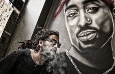 man with dreadlocks and sunglasses poses near Tupac Shakur portrait