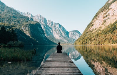 man sitting on gray dock looking out over a lake and mountains