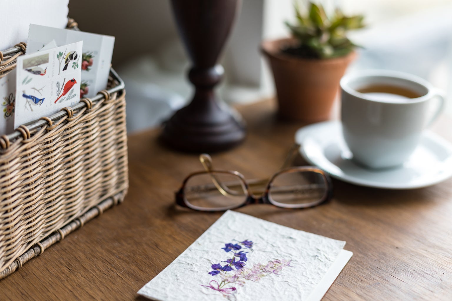white ceramic mug beside eyeglasses, card with print of purple flowers, and basket of postage stamps