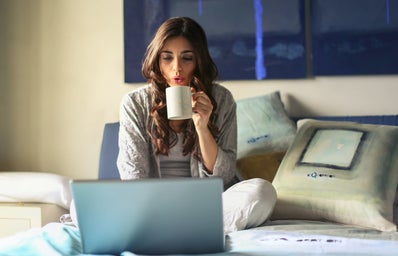 woman sitting on a bed in a gray cardigan drinking coffee and using her computer