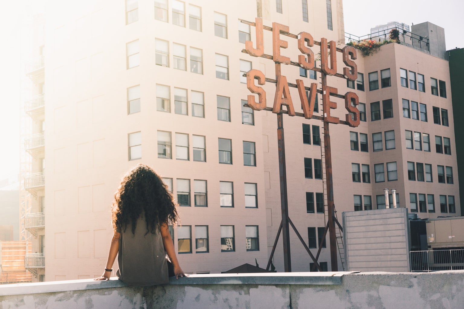 woman in gray top sitting on a building\'s edge in front of \"Jesus Saves\" sign