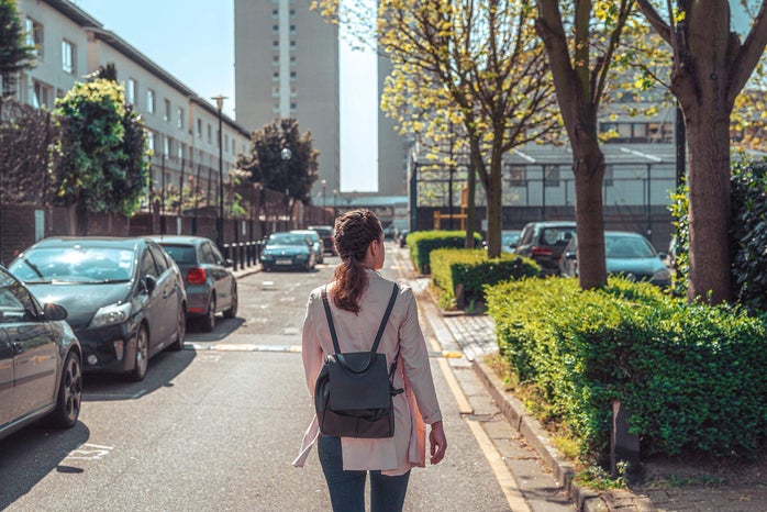 Photo of young brunette woman wearing a backpack and walking down a street alone shot from behind