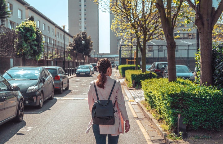 Photo of young brunette woman wearing a backpack and walking down a street alone shot from behind