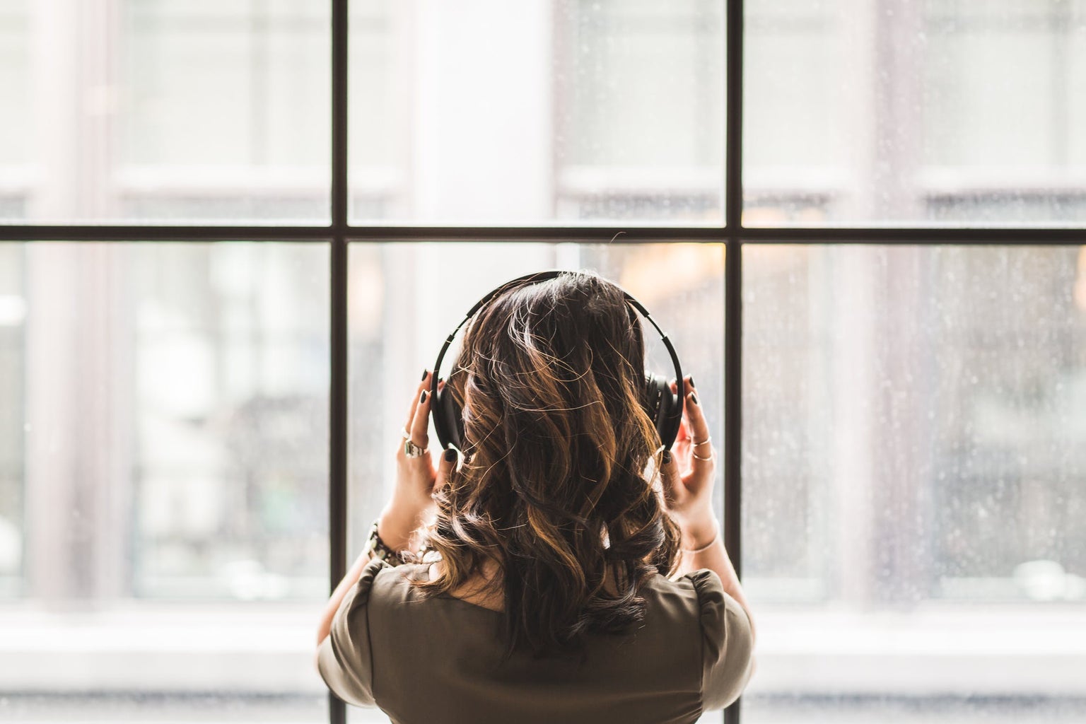 brunette woman shot from behind facing a window, wearing headphones listening to music and holding them with her hands
