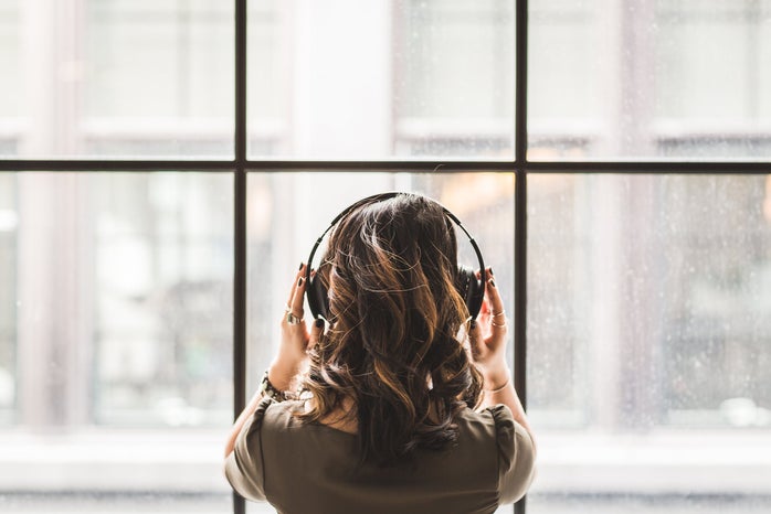 brunette woman shot from behind facing a window, wearing headphones listening to music and holding them with her hands
