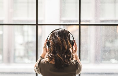 brunette woman shot from behind facing a window, wearing headphones listening to music and holding them with her hands