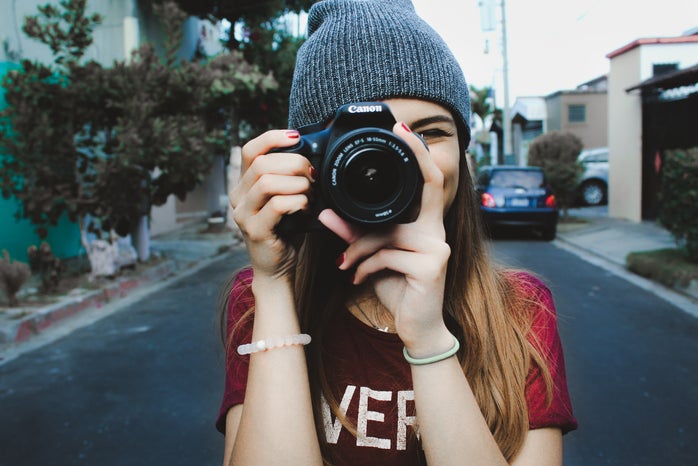 woman wearing red t-shirt and gray knitted cap standing on concrete road using Canon bridge camera during daytime