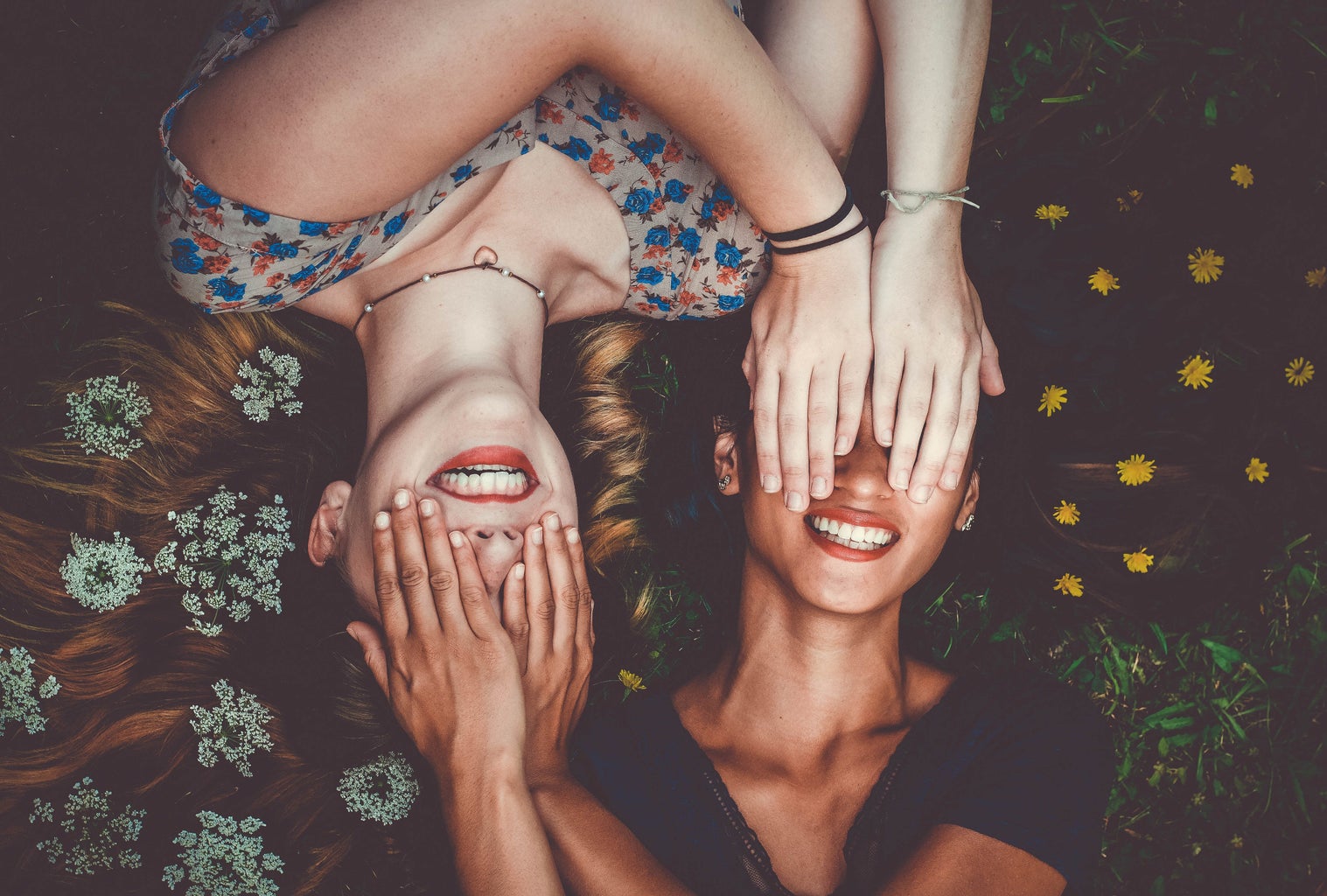 two woman smiling while laying on lawn field and covering each other\'s eyes