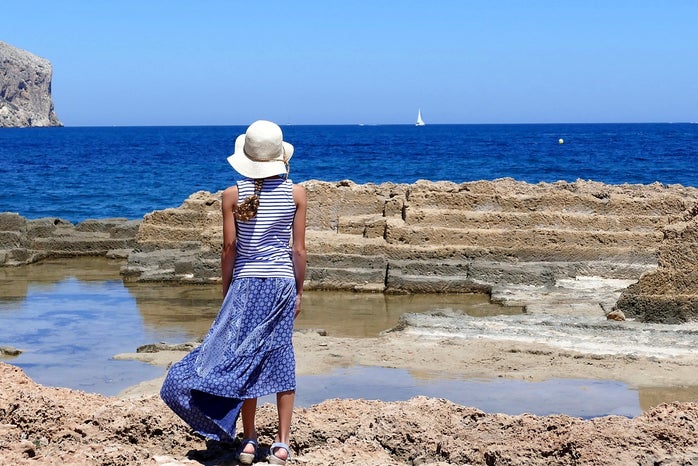woman in blue dress and white hat facing the ocean