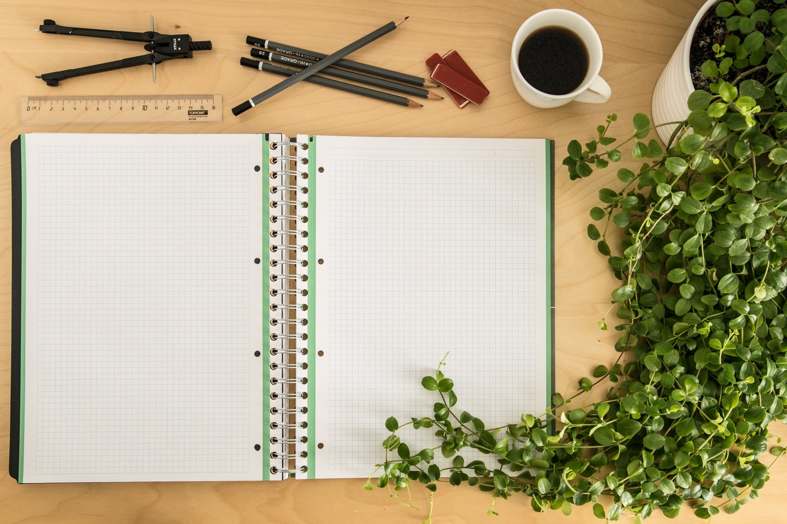 green and white grid notebook on desk with pencils, coffee, and plant