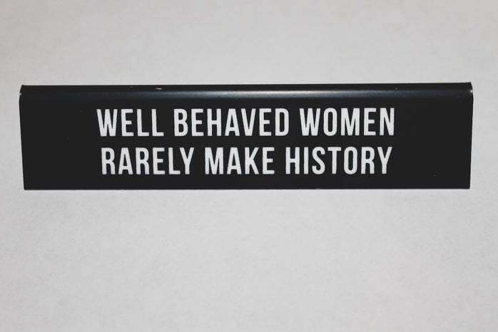 black placard stating on \"WELL BEHAVED WOMEN RARELY MAKE HISTORY\" on white table