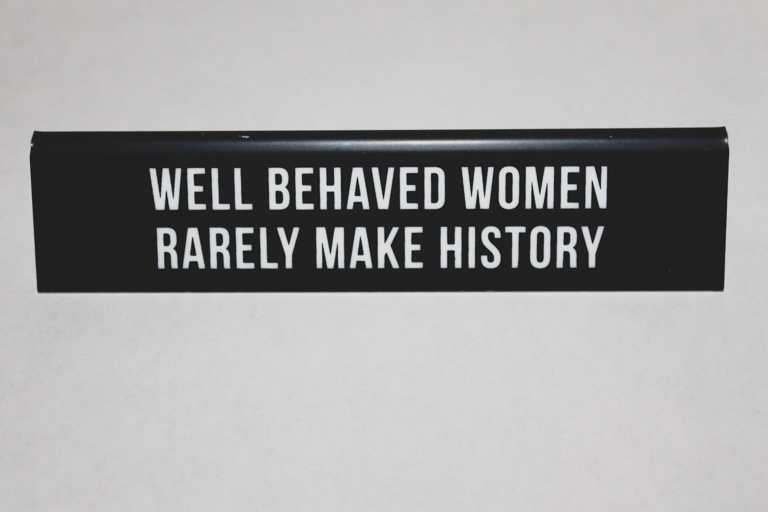 black placard stating on \"WELL BEHAVED WOMEN RARELY MAKE HISTORY\" on white table