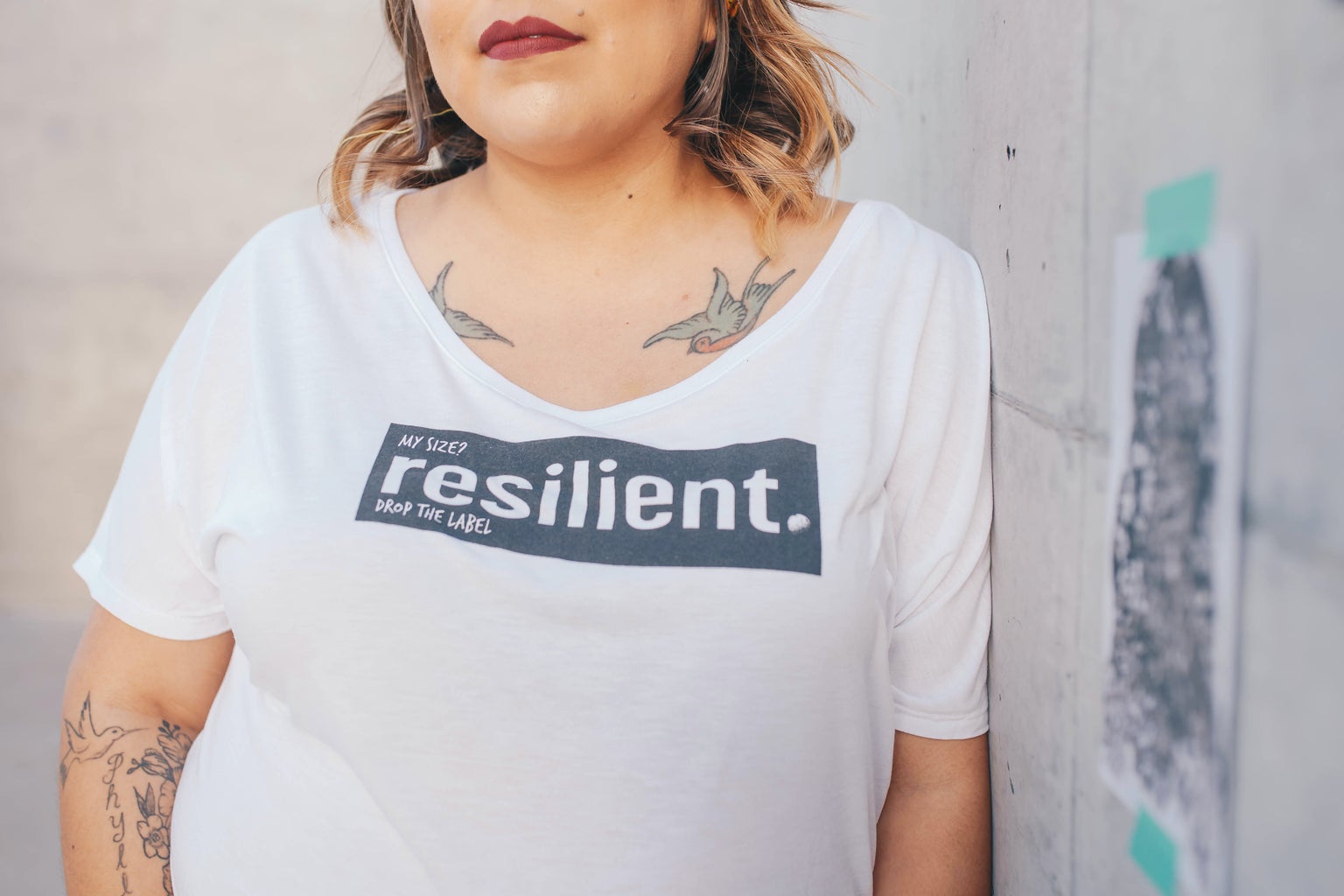 woman leaning on white wall wearing a white tee with text \