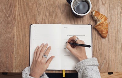 A person writing in a journal on a wooden table with a coffee cup and a croissant
