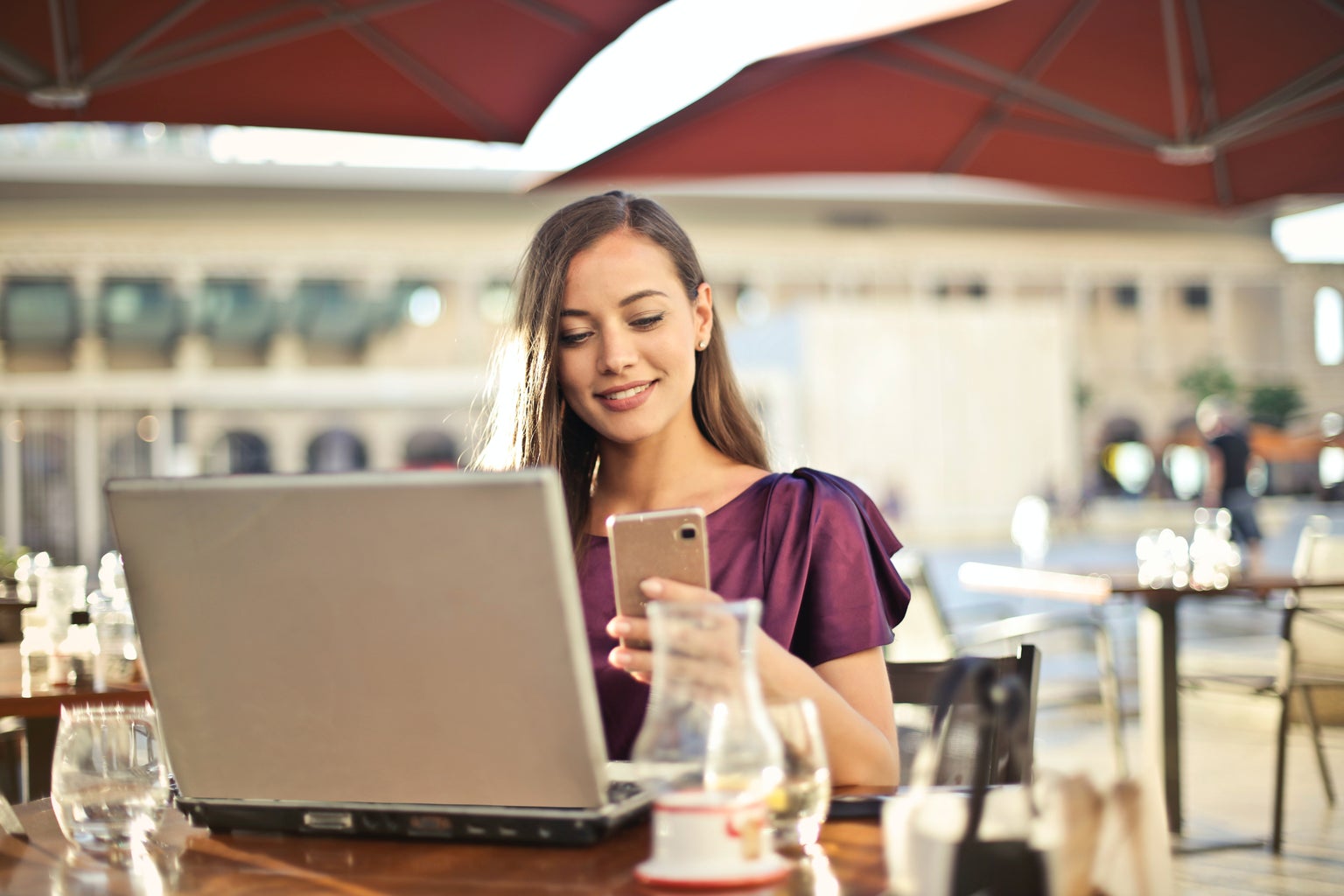 Woman sitting at a restaurant table outside on her phone with a laptop on the table in front of her.