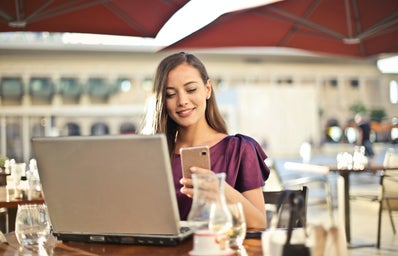 Woman sitting at a restaurant table outside on her phone with a laptop on the table in front of her.