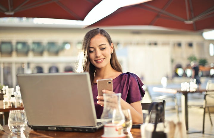 Woman sitting at a restaurant table outside on her phone with a laptop on the table in front of her.