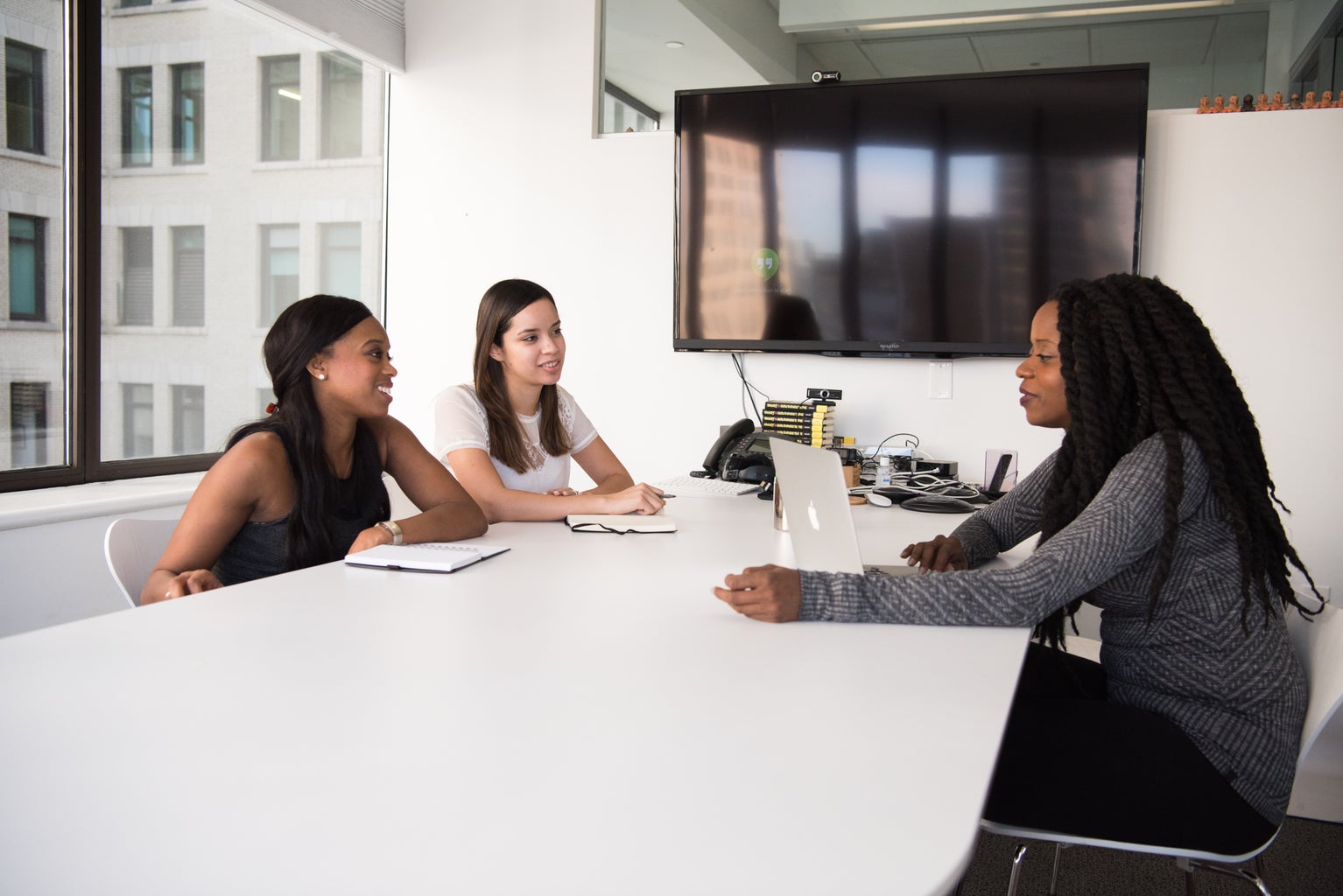 Three women of color are sitting at a table; two are on one side and  one is on the other with a laptop in front of her. They are in a conference room.