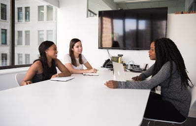 Three women of color are sitting at a table; two are on one side and  one is on the other with a laptop in front of her. They are in a conference room.