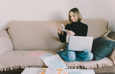 Woman with chin-length hair and a black turtleneck sits on a couch with her laptop and papers.